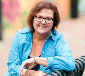 Portrait of HelenKay in a blue button down shirt and glasses sitting on a park bench